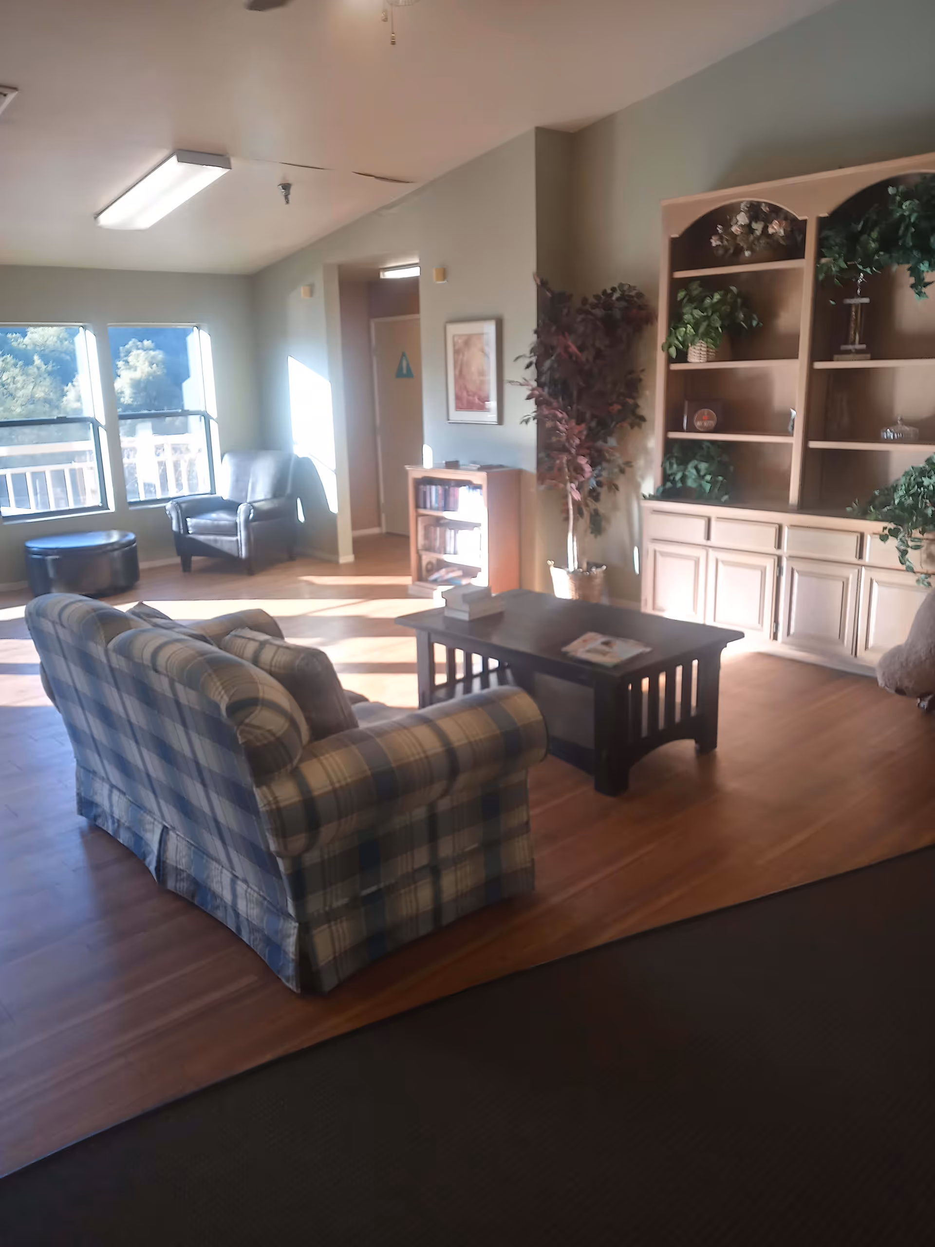 A cozy living room area in a senior living facility with a plaid sofa facing a dark wooden coffee table. There is a leather armchair and a round ottoman near two large windows letting in natural light. A tall wooden bookshelf with plants and decorative items is against the wall, and a potted plant stands next to it. The floor is wood, and there is a small bookshelf near a doorway with a restroom sign.