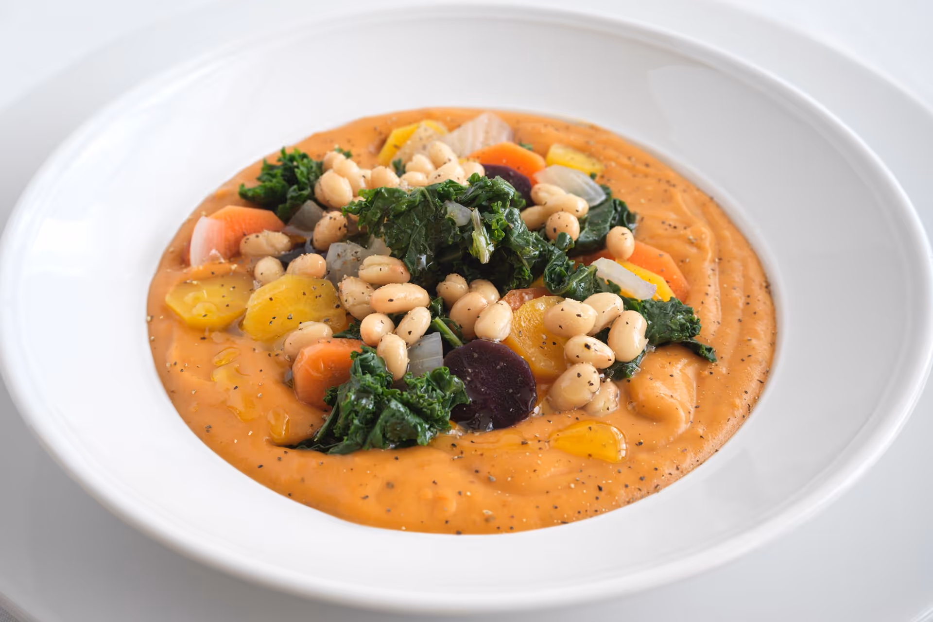 A white bowl filled with creamy orange soup topped with white beans, kale, and sliced colorful vegetables including carrots and beets, garnished with black pepper and a drizzle of oil.