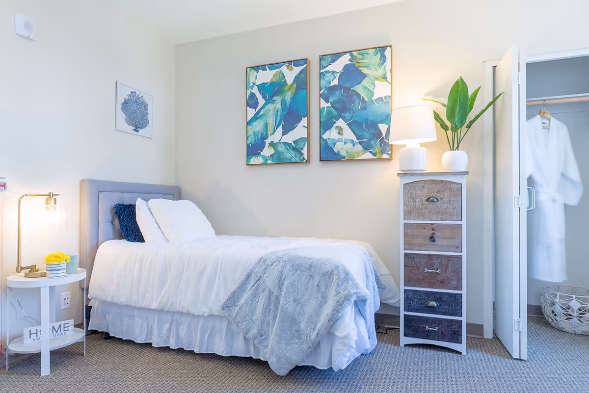 A neatly made single bed with white bedding and a gray throw blanket in a bright bedroom. Next to the bed is a small white round side table with a lamp, a small plant, and a decorative sign that says 'HOME SWEET HOME'. On the wall above the bed are two framed artworks featuring blue and green tropical leaves. To the right of the bed is a tall chest of drawers with a white lamp and a potted plant on top. An open closet door reveals a white robe hanging inside and a woven basket on the floor.