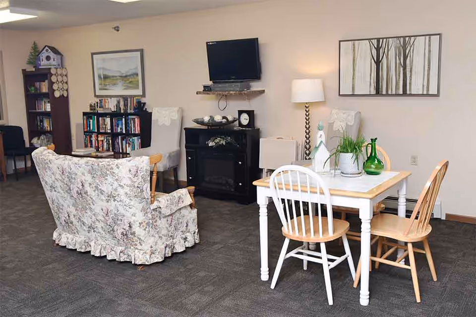 A cozy living area with a floral patterned sofa, two upholstered chairs, a small wooden dining table with four chairs, and a TV mounted on the wall above a black cabinet. The room also features a bookshelf filled with books, a floor lamp, and framed artwork on the walls.