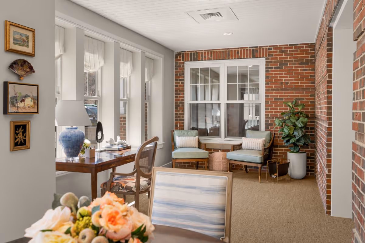A cozy sitting area in a senior living facility with two light blue cushioned chairs, a small round wicker table, a potted plant, and a wooden desk with a blue lamp and decorative items. The room features large windows with white curtains, exposed brick walls, and framed artwork on a gray wall.