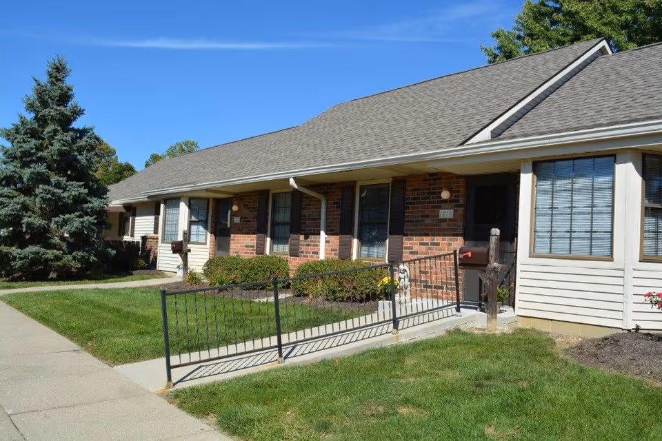 Exterior view of a single-story residential building with a brick and siding facade, multiple windows, a front door, a mailbox, a ramp with a black metal railing, green grass, and a large evergreen tree under a clear blue sky.