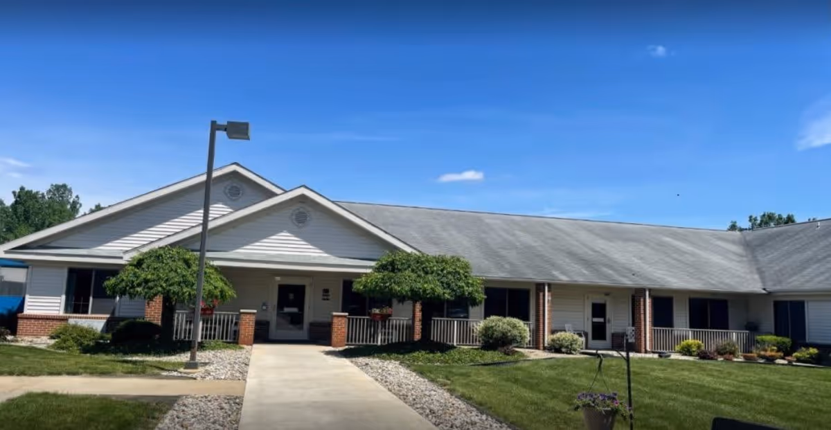 Exterior view of a single-story assisted living facility building with a gray roof, white siding, and brick accents. There is a concrete walkway leading to the entrance, flanked by small trees and landscaped bushes under a clear blue sky.