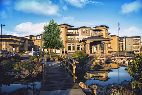 Exterior view of a multi-story senior living facility named Bonaventure of Salem with a wooden footbridge over a pond in the foreground, landscaped greenery, and a clear blue sky.