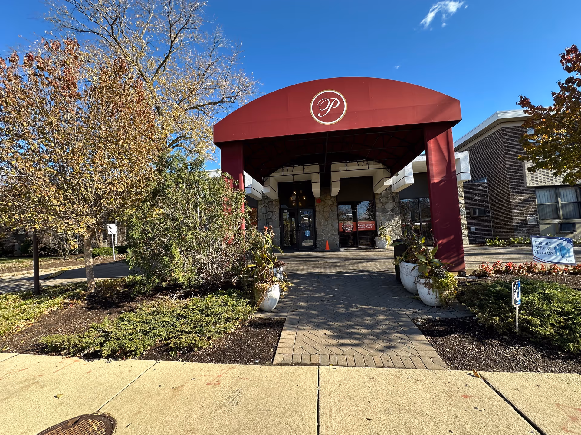 Entrance of Peterson Park Health Care Center with a red canopy featuring a circular emblem with the letter 'P'. The building has stone and brick exterior walls, surrounded by landscaped bushes and trees with autumn foliage under a clear blue sky.