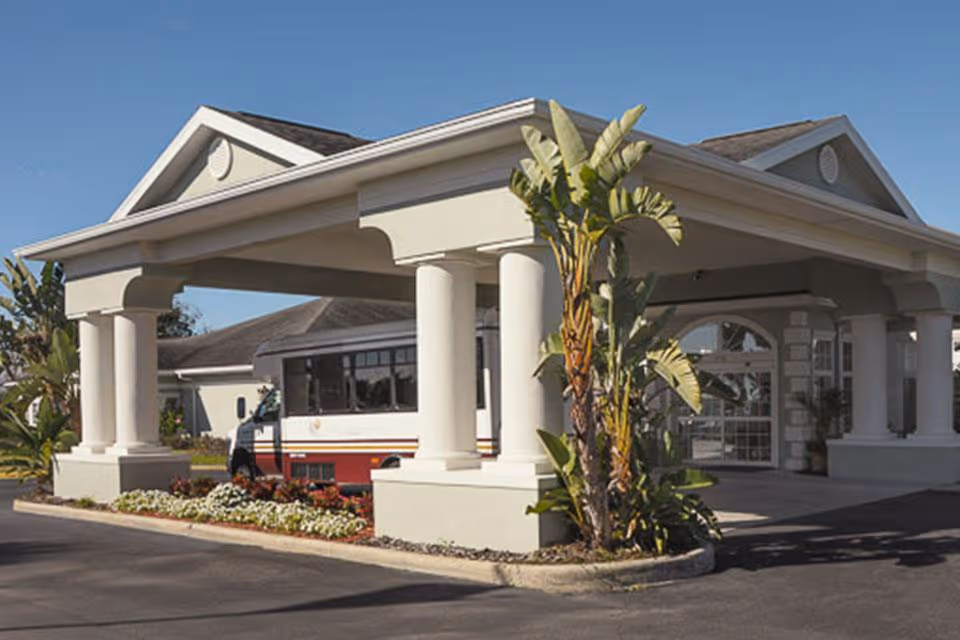 Front exterior view of a senior living facility with a covered entrance supported by large white columns, landscaped with plants and flowers, and a shuttle bus parked nearby under a clear blue sky.