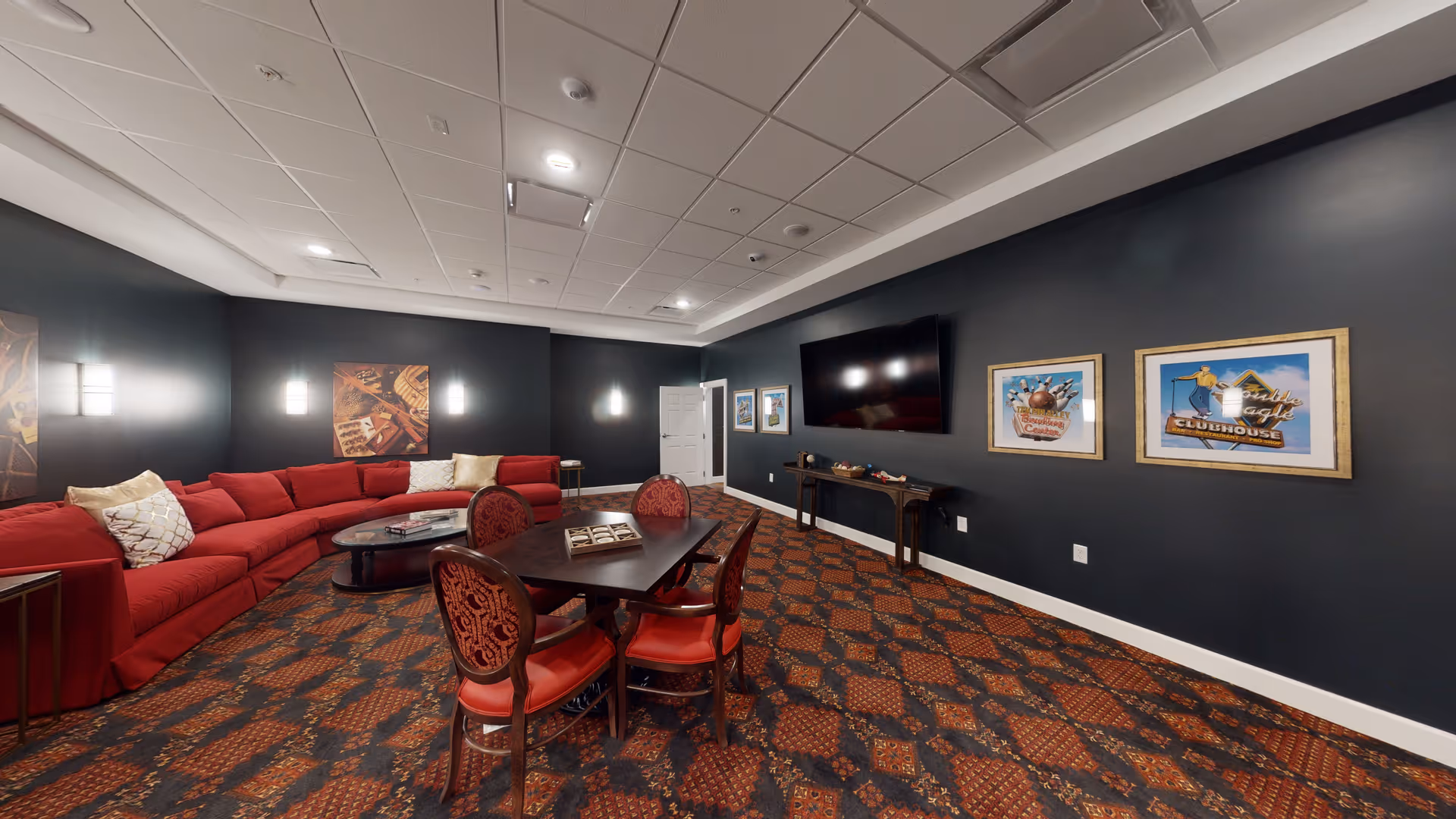 A cozy senior living common area with a large red sectional sofa, a dark wood coffee table, and a dining table with four red upholstered chairs. The walls are painted dark gray and decorated with framed pictures. A large flat-screen TV is mounted on one wall above a narrow console table. The carpet has a patterned design in red and dark tones, and the ceiling has recessed lighting.