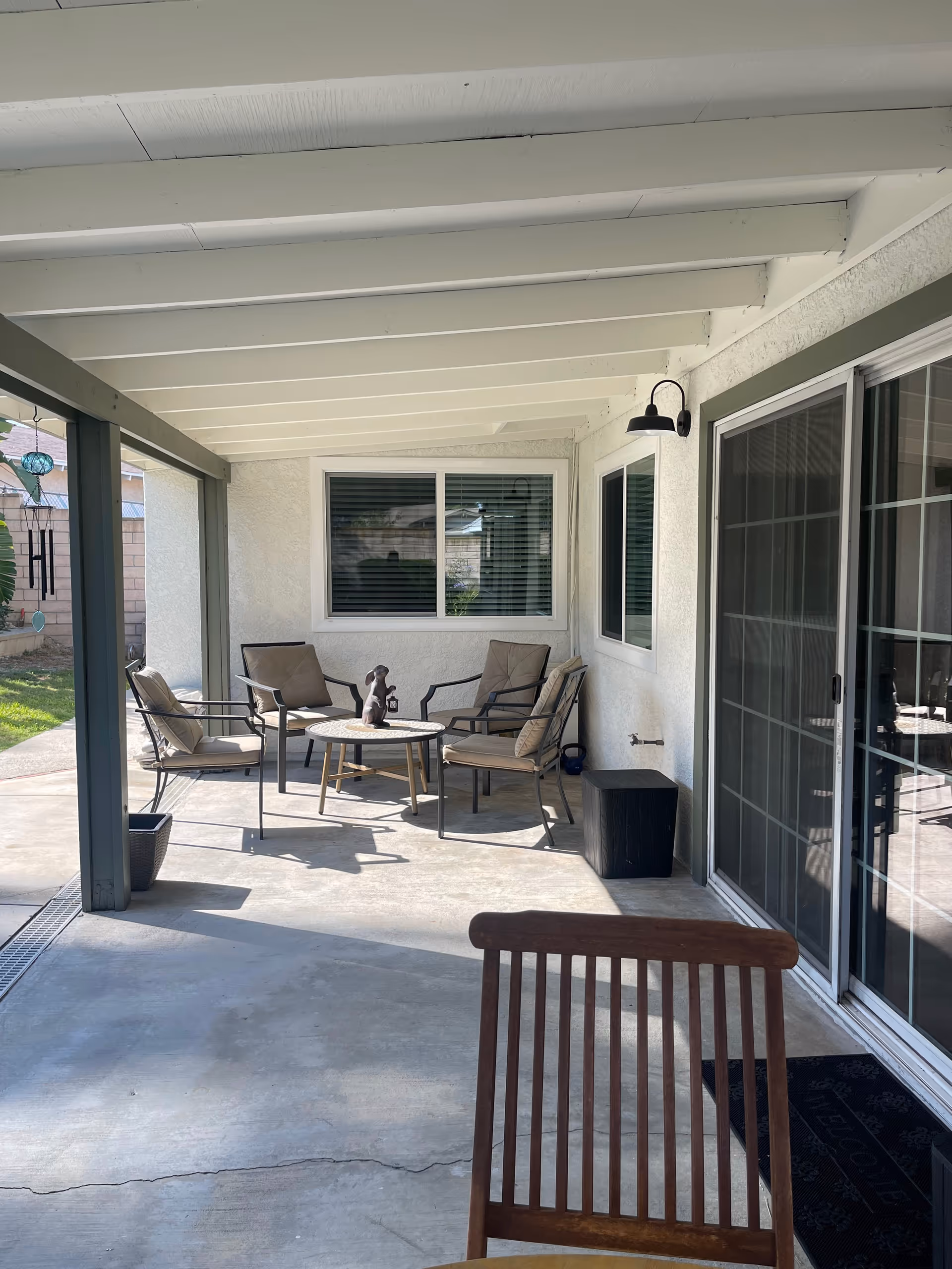 Covered outdoor patio area with a concrete floor, four cushioned chairs arranged around a small round table with a decorative sculpture on it. The patio has white beams overhead, a sliding glass door on the right, and a window with blinds on the back wall. A wooden chair is partially visible in the foreground.