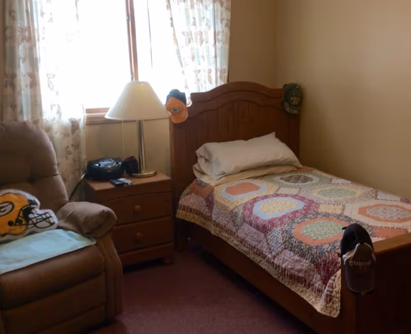 A cozy bedroom with a single wooden bed covered with a colorful patchwork quilt. There are two pillows on the bed. A wooden nightstand beside the bed holds a lamp and a small radio. A comfortable armchair with a Green Bay Packers helmet pillow is next to the nightstand. The room has a window with floral curtains letting in natural light. Several baseball caps are hanging on the bed's headboard and footboard.