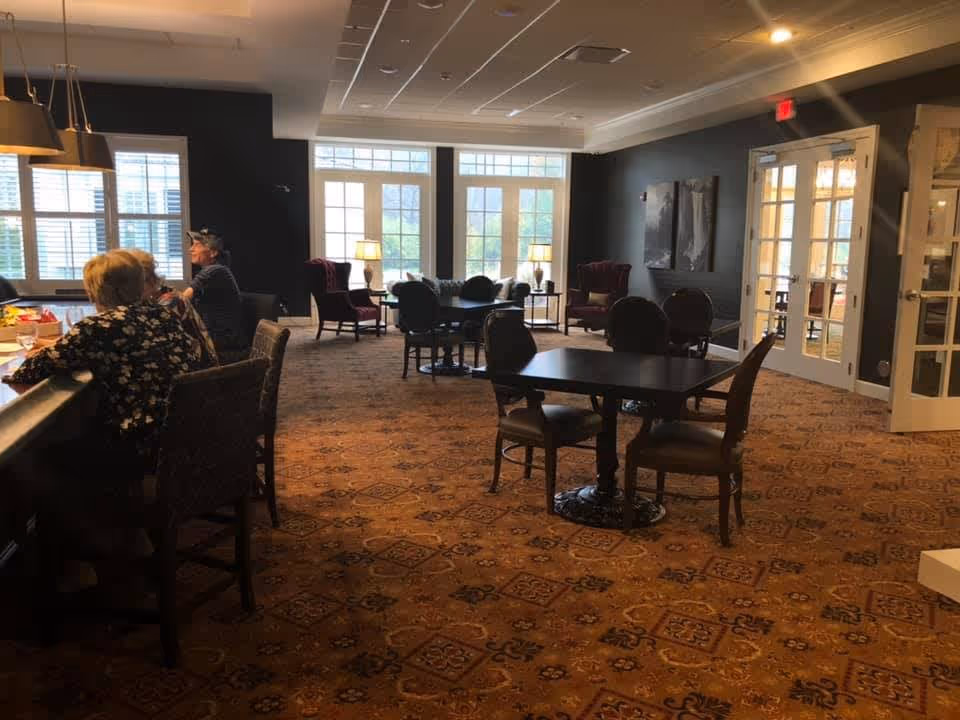 A spacious common area in a senior living facility with patterned carpet, several tables and chairs, and large windows letting in natural light. Two elderly people are seated at a counter on the left side, and there are armchairs and lamps near the windows. Double glass doors are visible on the right side of the room.
