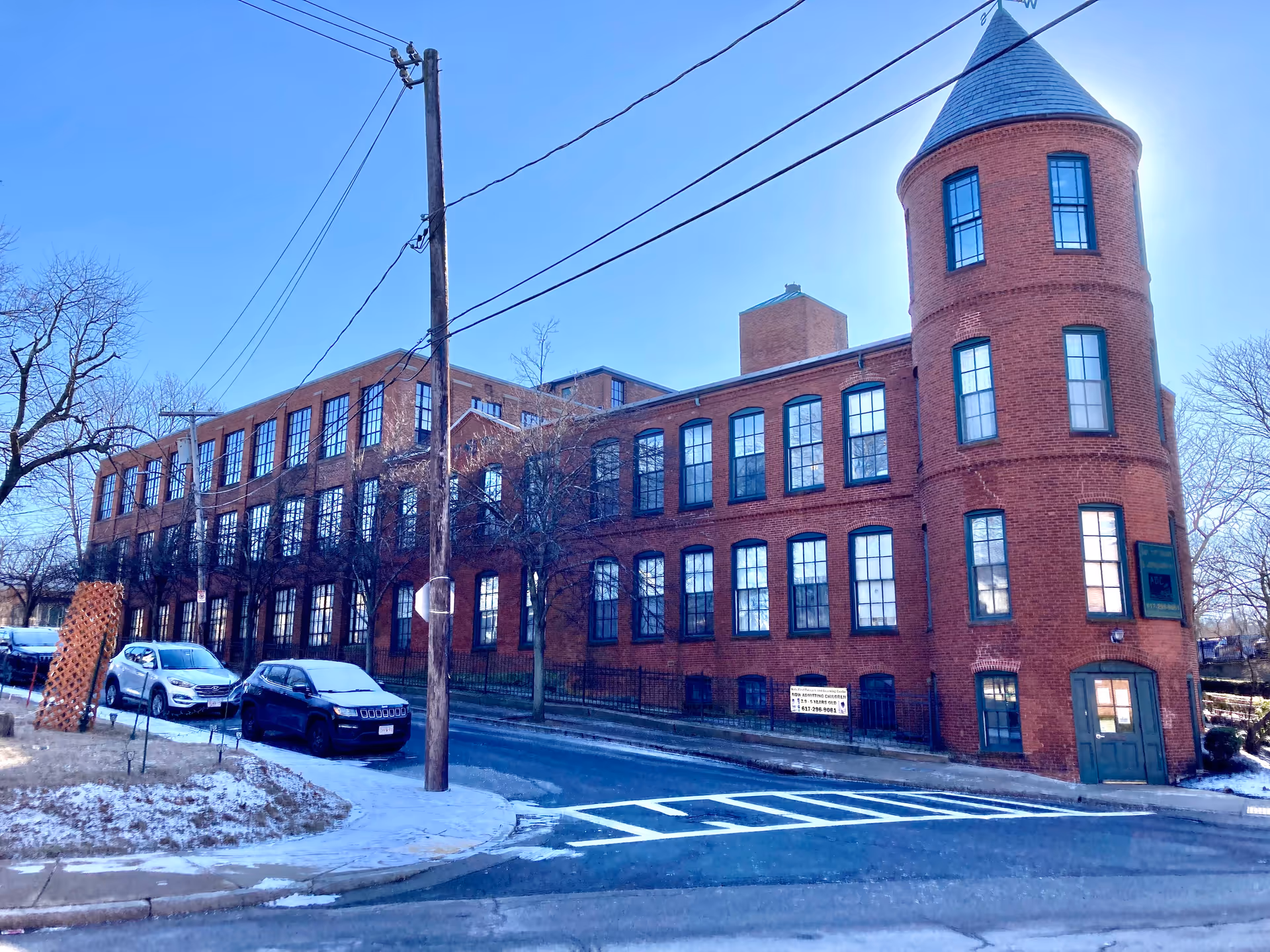 Exterior view of a large red brick building with many windows and a rounded turret on the right side. There are a few parked cars along the street in front of the building, and some snow on the ground. The sky is clear and blue.
