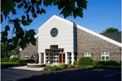 Exterior view of a single-story building with a white triangular front facade featuring a circular window. The building has a combination of white siding and brick walls, surrounded by green shrubs and trees under a clear blue sky.