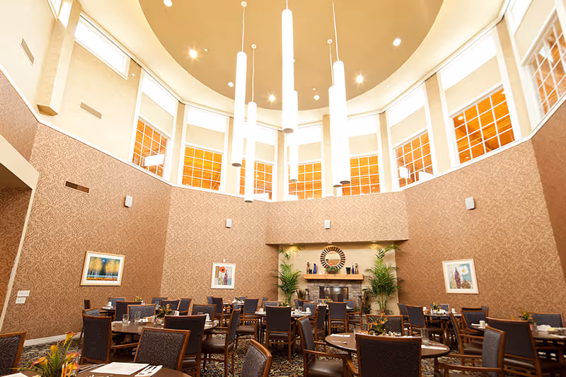 A spacious dining room with multiple round tables and chairs arranged neatly. The walls are decorated with patterned wallpaper and framed artwork. Tall windows near the ceiling allow natural light to fill the room, and modern pendant lights hang from the high ceiling. There are plants and decorative items on a mantelpiece at the far end of the room.
