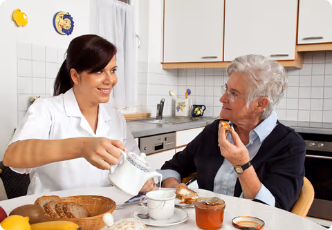 A young female caregiver in a white uniform pours tea from a teapot into a cup while sitting at a kitchen table with an elderly woman who is holding a piece of bread with jam. The table has a basket of bread, a jar of jam, and other breakfast items. The background shows a kitchen with white cabinets and tiled walls.