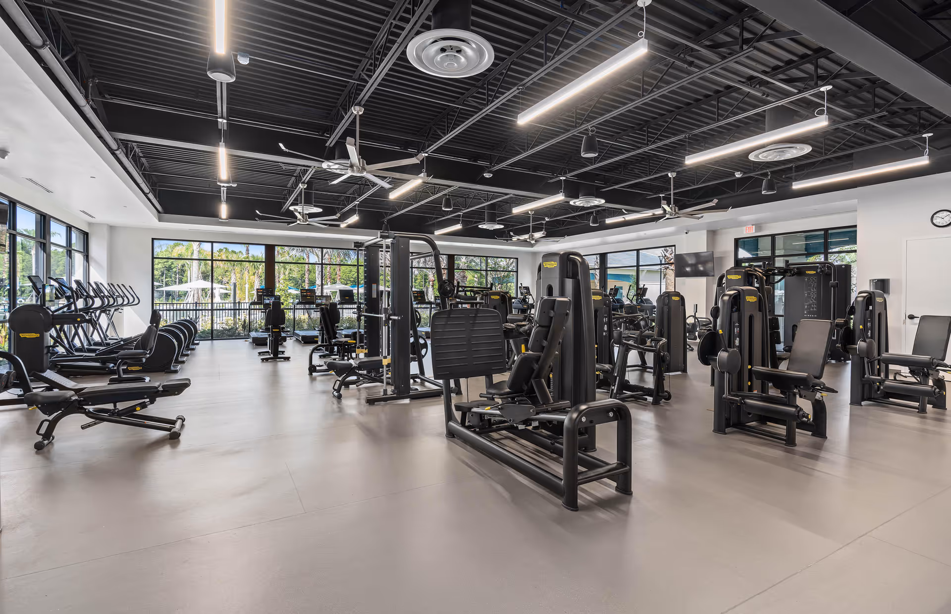 A spacious and modern fitness center with various exercise machines including treadmills, stationary bikes, and strength training equipment. The room has large windows letting in natural light and a high ceiling with exposed black beams and ceiling fans.