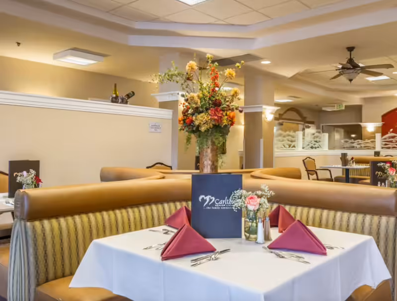 Dining area in Carlton Senior Living San Jose with a table set for four, featuring folded red napkins, silverware, a small flower arrangement, and a menu. The seating includes cushioned booths with striped upholstery, and a large floral centerpiece is visible in the background under warm lighting.
