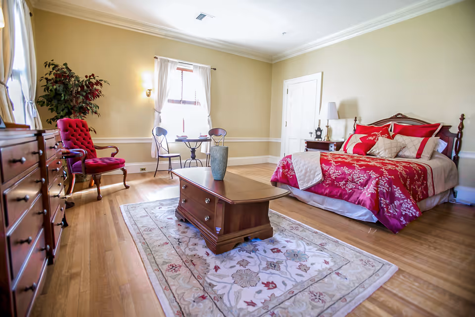 A bright bedroom with wooden flooring featuring a large bed with red and white floral bedding, a wooden coffee table on a patterned rug, a red upholstered armchair, a small round table with two chairs near a window with white curtains, and a wooden dresser. The walls are painted light beige with white trim.