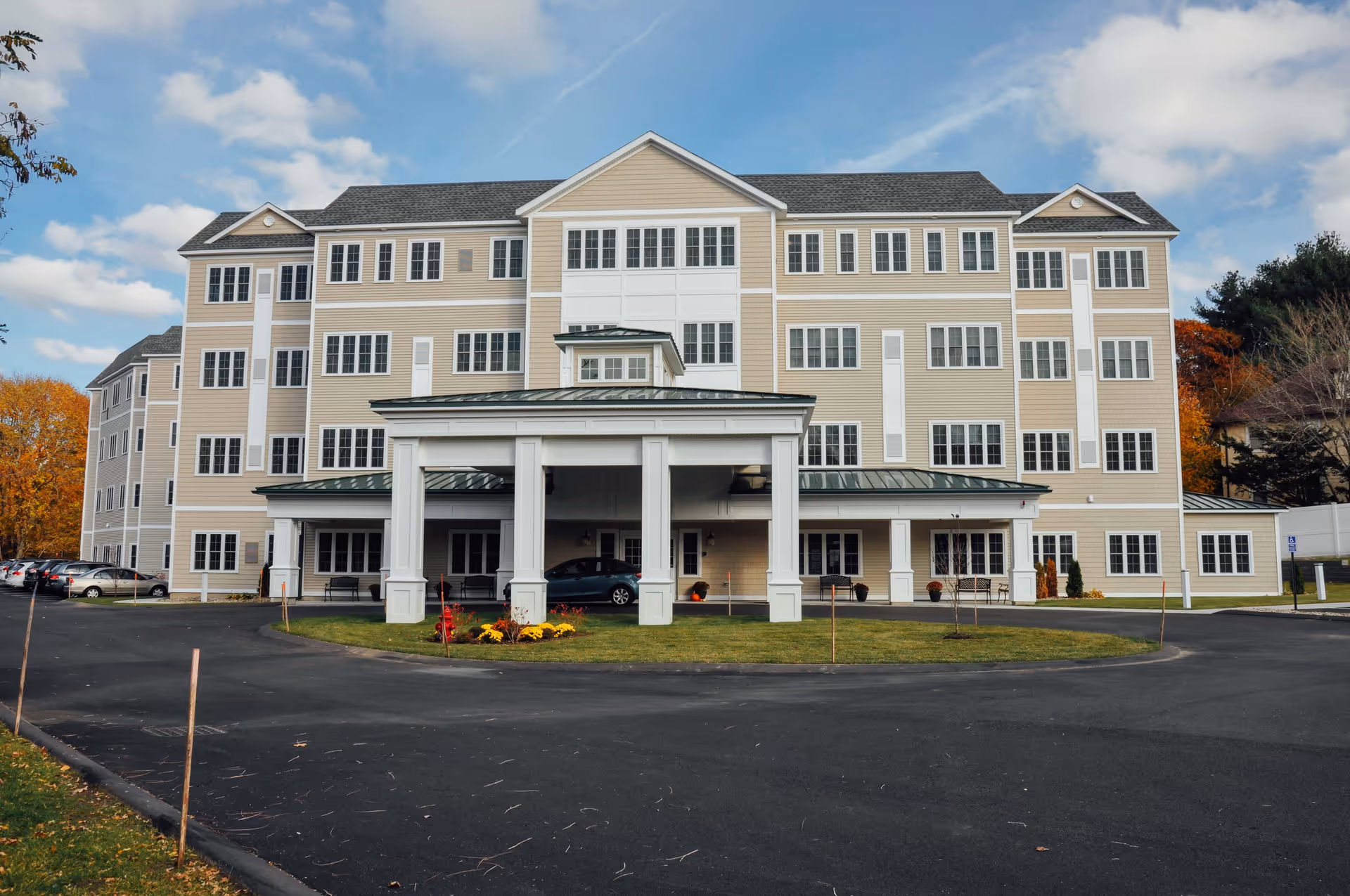 Front exterior view of a large, multi-story assisted living facility building with beige siding and numerous windows. The building has a covered entrance with white pillars and a circular driveway. There are some cars parked on the left side and autumn trees in the background under a partly cloudy sky.