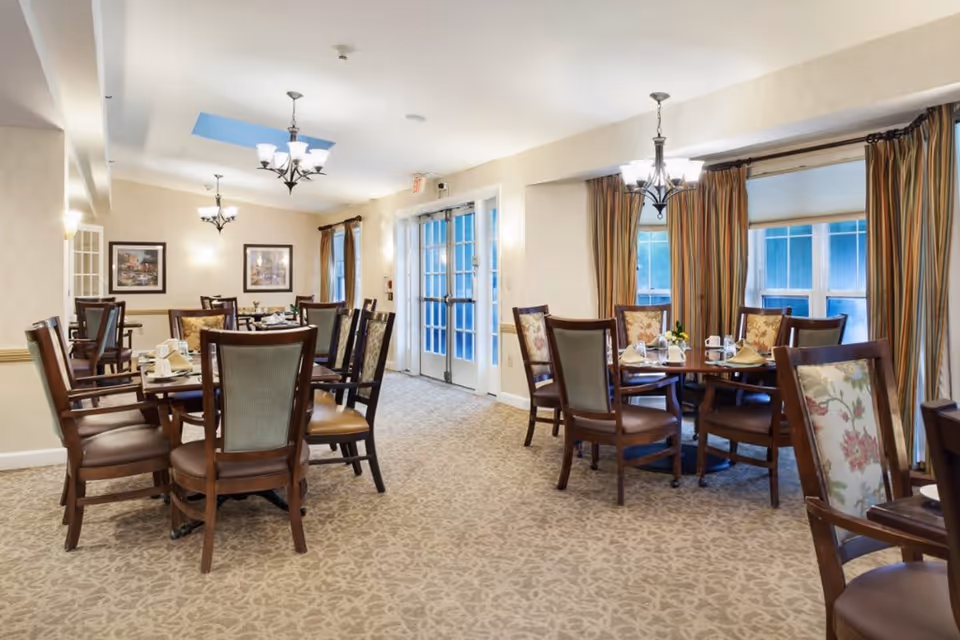 A well-lit dining room with multiple round tables set with napkins, cups, and small flower arrangements. The room features patterned carpet, wooden chairs with cushioned seats and backs, large windows with striped curtains, and chandeliers hanging from the ceiling. There are framed pictures on the walls and double glass doors at the far end of the room.
