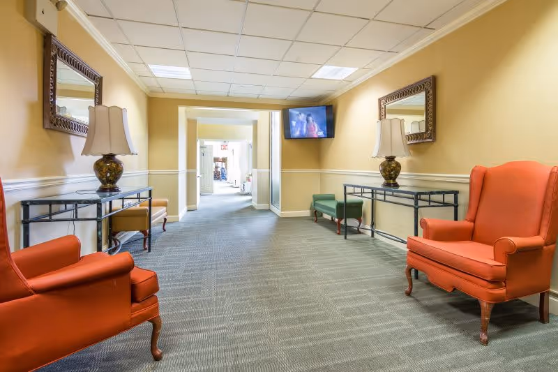 A hallway in a senior living facility with beige walls and carpeted floor. The hallway features two orange armchairs on either side, two console tables with decorative lamps and mirrors above them, a green bench, and a wall-mounted television. The hallway leads to a brighter area with open doors at the far end.