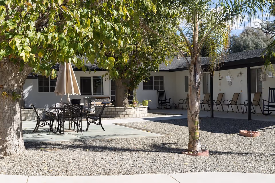 Outdoor courtyard area at Yorkshire Village featuring a seating area with a metal table and chairs under a large tree with an umbrella. The courtyard is surrounded by a single-story building with a covered porch lined with chairs and rocking chairs. There are palm trees and gravel ground covering.