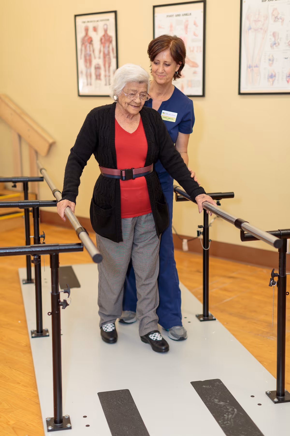 An elderly woman wearing a red shirt and black cardigan is walking between parallel bars for physical therapy, assisted by a healthcare worker in blue scrubs. The setting appears to be a rehabilitation or therapy room with anatomical charts on the wall.