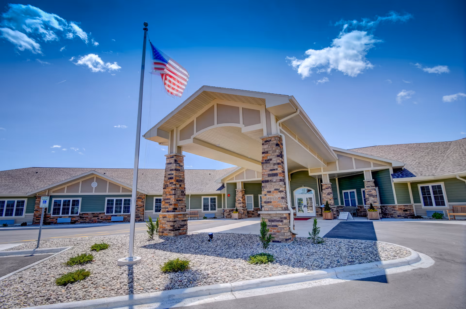 Front exterior view of The Courtyard at Bellevue Memory Care and Assisted Living facility with a covered entrance supported by stone pillars, an American flag on a flagpole, and a clear blue sky with some clouds.