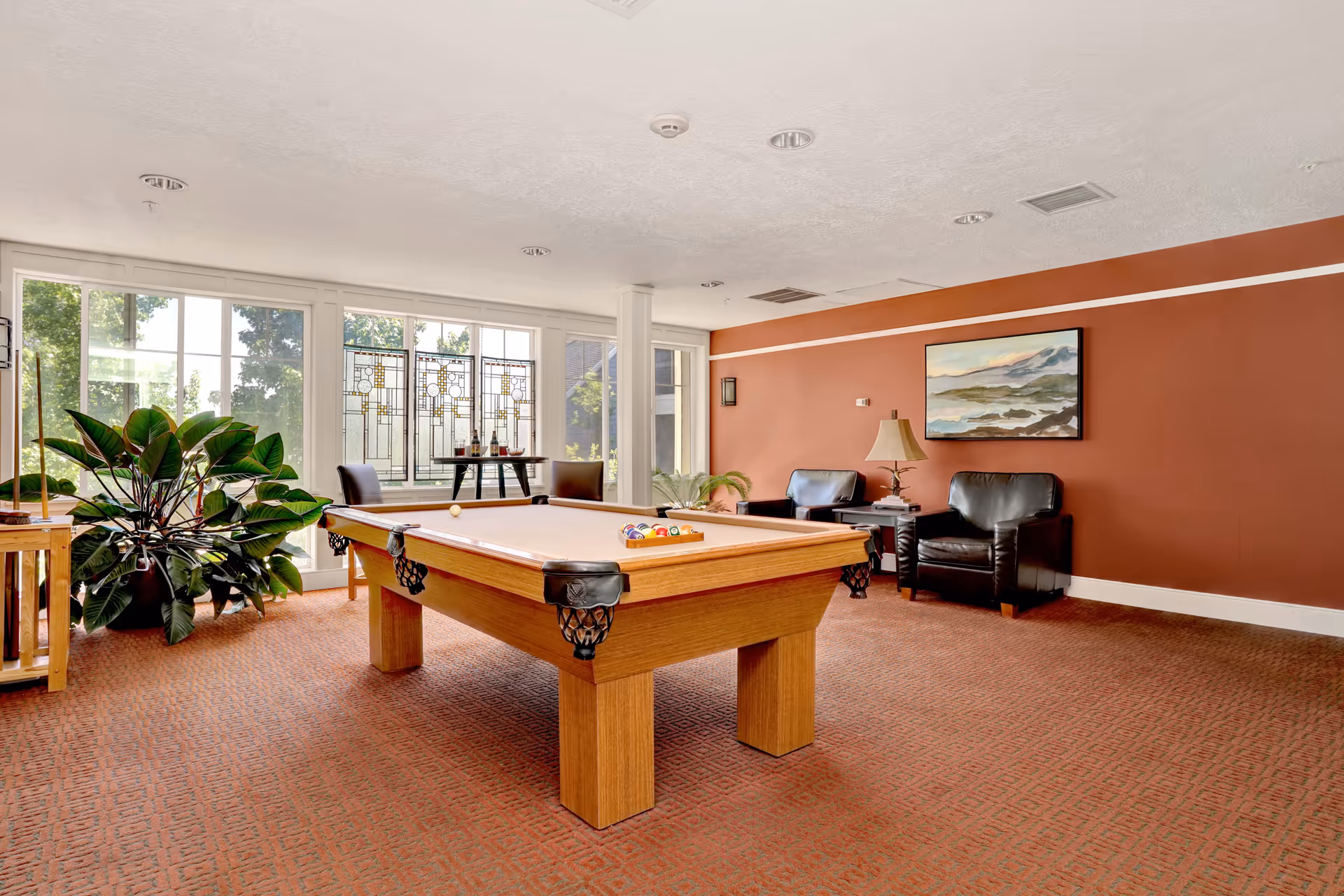 A well-lit game room with a pool table in the center, two black leather armchairs against a rust-colored wall, a small table with a lamp between the chairs, a large plant near the window, and a painting hanging on the wall. Large windows allow natural light to fill the room.