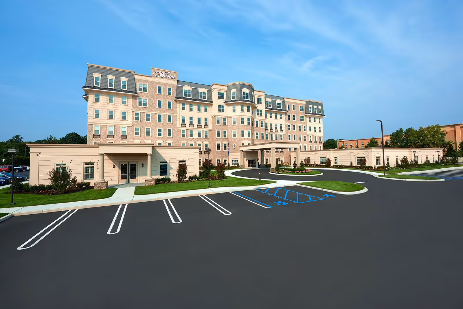 Exterior view of The Bristal at Garden City, a multi-story senior living facility with a large parking lot in front, landscaped greenery, and a clear blue sky.