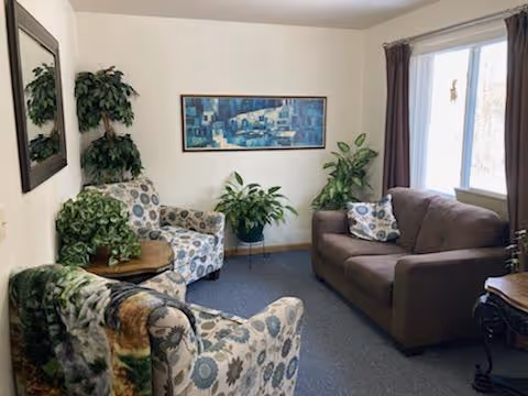 A cozy living room with a brown sofa and two patterned armchairs. There are several green plants placed around the room, a large window with brown curtains letting in natural light, a rectangular framed artwork on the wall, and a mirror reflecting part of the room.