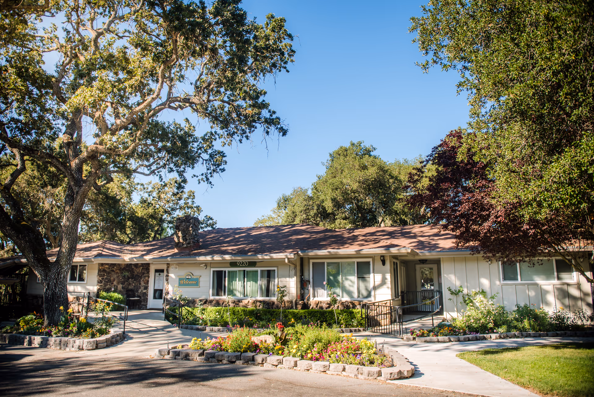 Single-story building with a brown shingled roof and white exterior walls, surrounded by large trees and colorful flower beds. A paved walkway with handrails leads to the entrance, which has a 'Welcome' sign above the door. The sky is clear and blue.