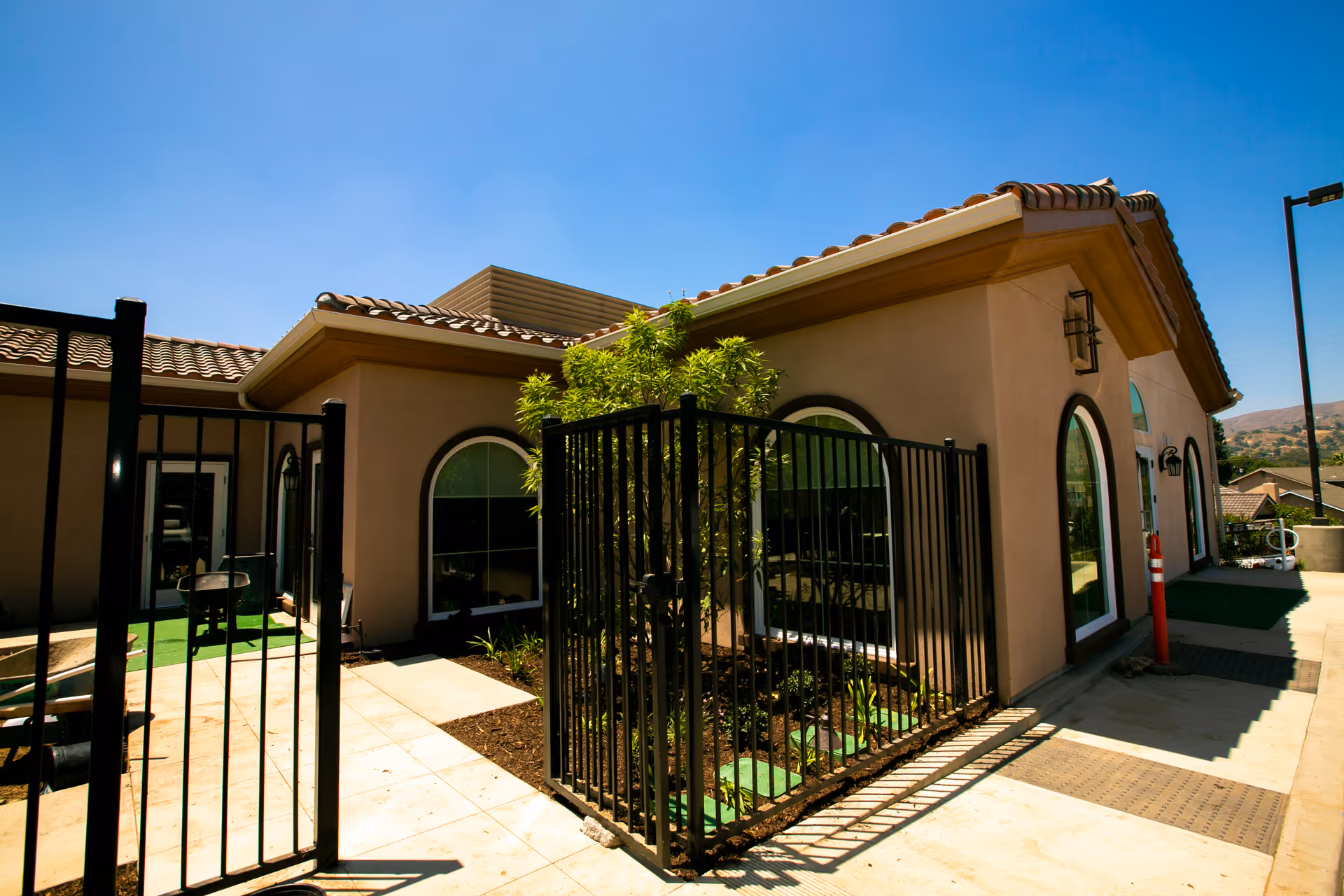 Exterior view of a single-story building with beige walls and a tiled roof under a clear blue sky. The building has arched windows and a black metal fence enclosing a small garden area with plants. A paved walkway surrounds the building.