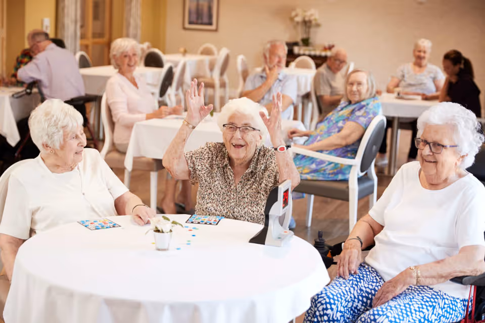 A group of elderly people sitting at round tables in a bright room, engaging in a game of bingo. Three elderly women are seated at the front table, one raising her hands excitedly, while others in the background are also seated and appear to be enjoying the activity.