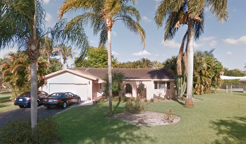 Single-story house with a tiled roof surrounded by palm trees and green lawn. Two cars are parked in the driveway. The sky is clear with a few clouds.