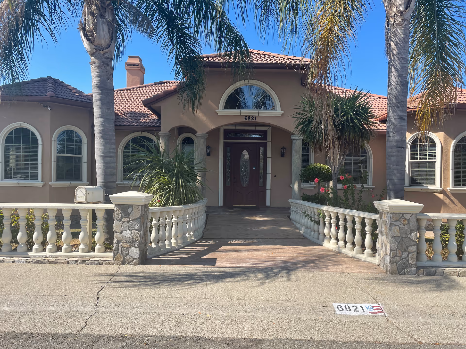 Front exterior view of a single-story building with a tiled roof, arched windows, and a central entrance door. The entrance is flanked by two palm trees and stone pillars with white balustrades on either side of the walkway. The building number 6821 is visible above the door and painted on the curb with an American flag next to it. The sky is clear and blue.