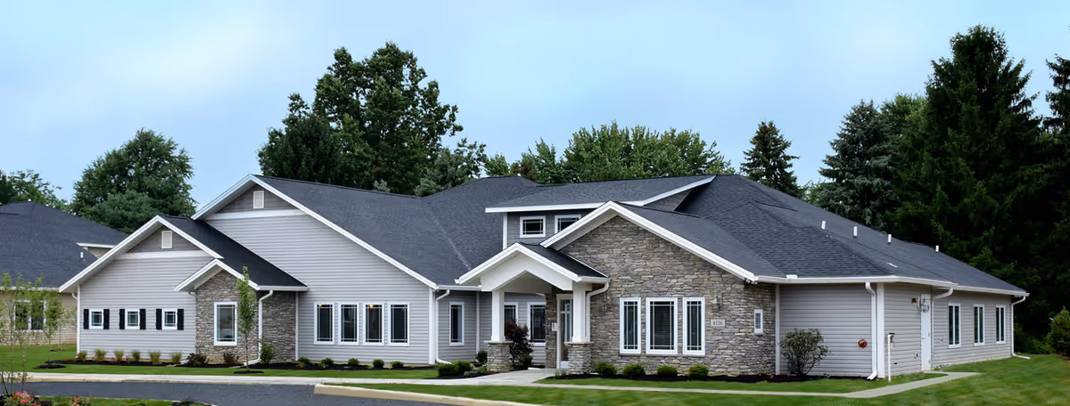Exterior view of a single-story senior living facility building with gray siding and stone accents, surrounded by green grass and trees under a cloudy sky.