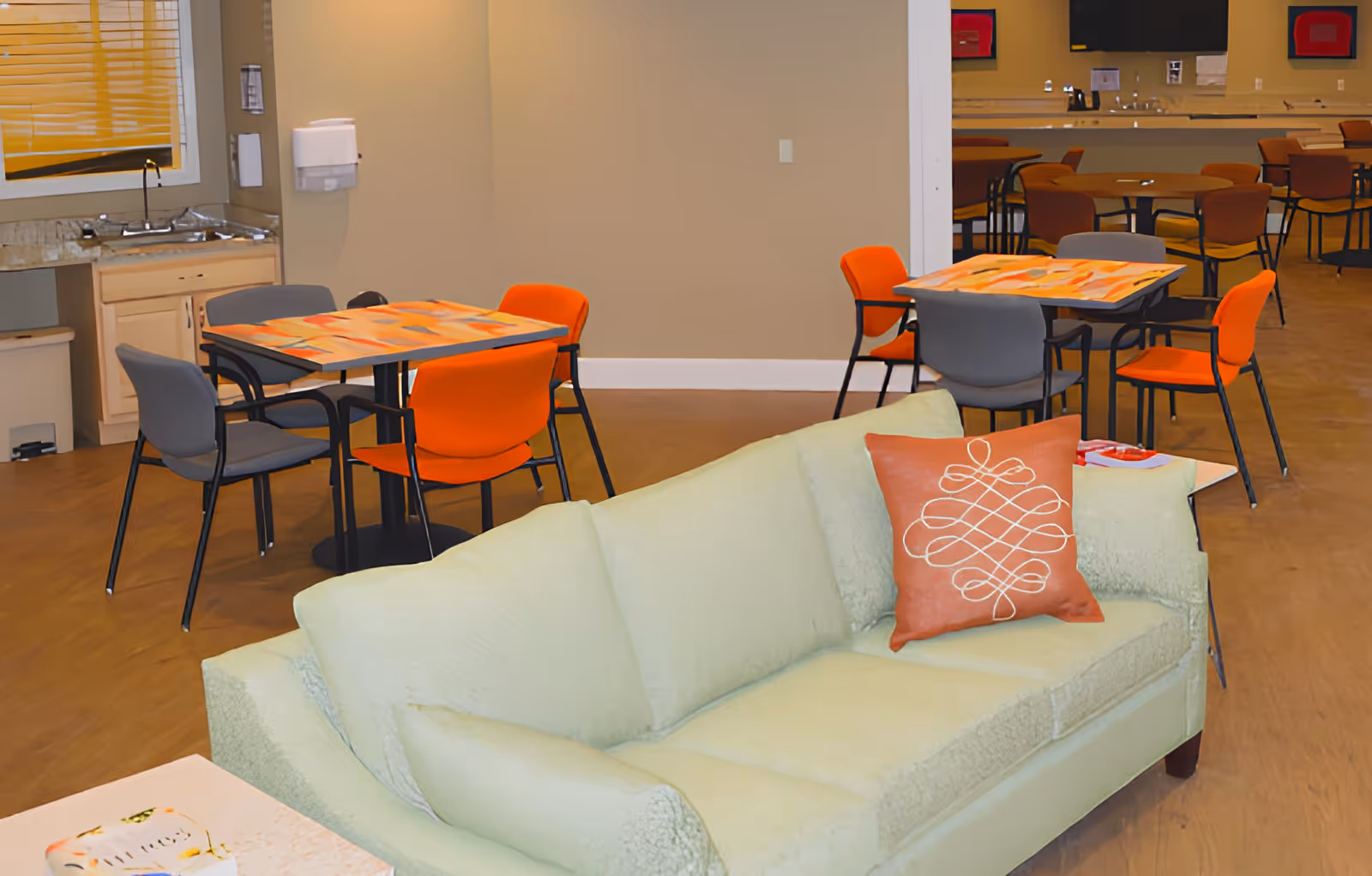 Interior view of a senior living facility common area with a light green sofa featuring an orange decorative pillow in the foreground. Behind the sofa are several tables with orange and gray chairs. A small kitchenette with a sink and countertop is visible on the left side of the image.