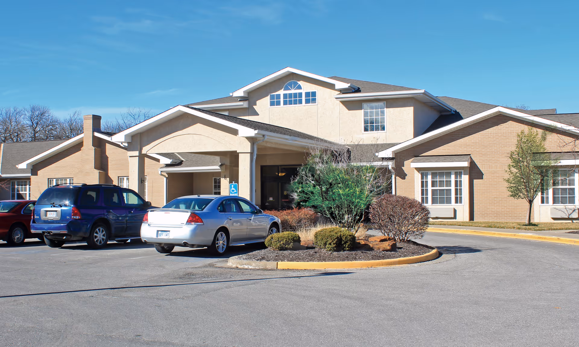 Exterior front view of a single-story senior living facility building with a covered entrance, several windows, and a small landscaped area with bushes and trees. There are three cars parked in front of the building under a clear blue sky.