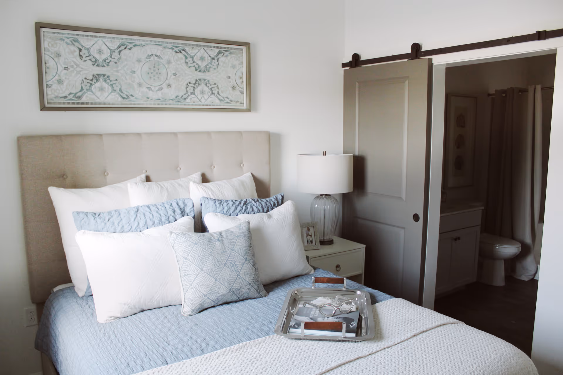 A neatly made bed with multiple white and light blue pillows and a light blue quilted bedspread. A silver tray with a pair of eyeglasses and a book rests on the bed. To the right, there is a nightstand with a glass lamp and a small framed photo. A sliding barn door is partially open, revealing a bathroom with a toilet, vanity, and shower curtain. A decorative framed artwork hangs above the bed on a white wall.