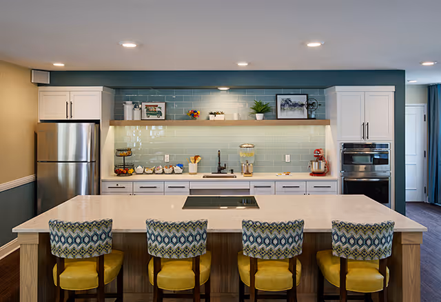Modern kitchen with a large island countertop featuring four patterned cushioned bar stools with yellow seats. The kitchen has white cabinets, a stainless steel refrigerator, built-in oven, and a light blue tiled backsplash. Open wooden shelves display decorative items and kitchen essentials.