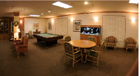 A cozy common room with a pool table in the center, a round table with four chairs, two armchairs against the wall, a large fish tank on a wooden cabinet, framed artwork on the walls, and warm lighting from wall sconces and a floor lamp.