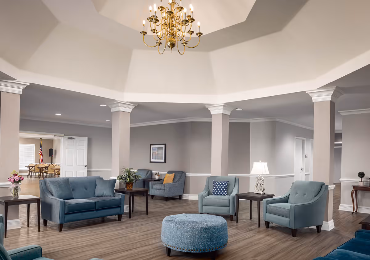 A spacious senior living facility common area with light gray walls, wooden flooring, and several blue upholstered chairs and sofas arranged around dark wooden side tables. A round blue ottoman is in the center. The ceiling features a large, elegant chandelier and white crown molding. In the background, there is a room with tables and chairs and an American flag.