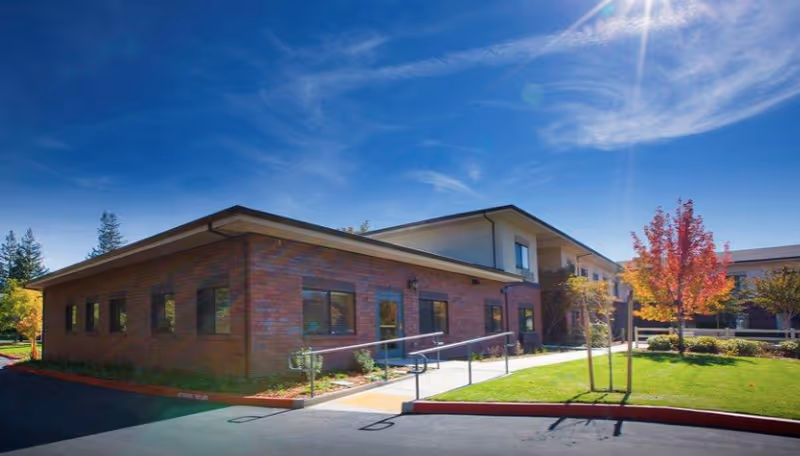 Exterior view of a two-story brick and stucco building under a bright blue sky with some wispy clouds. The building is surrounded by a well-maintained lawn, a few small trees including one with red autumn leaves, and a paved driveway with a wheelchair ramp leading to the entrance.