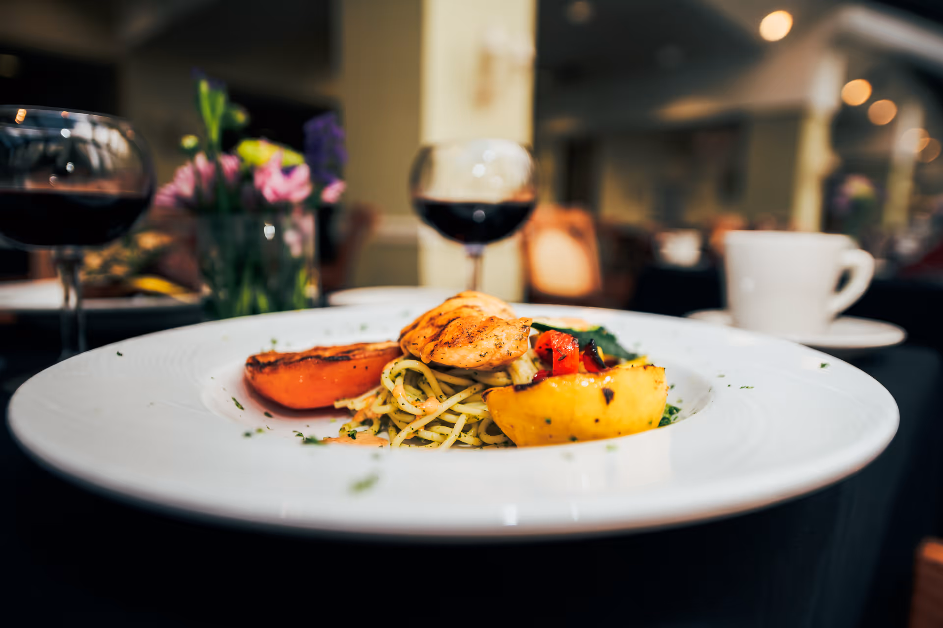 Close-up of a gourmet meal featuring grilled chicken on a bed of pasta with grilled vegetables, served on a white plate. In the background, there are two glasses of red wine, a white coffee cup, and a vase with flowers on a table in a softly lit dining room.