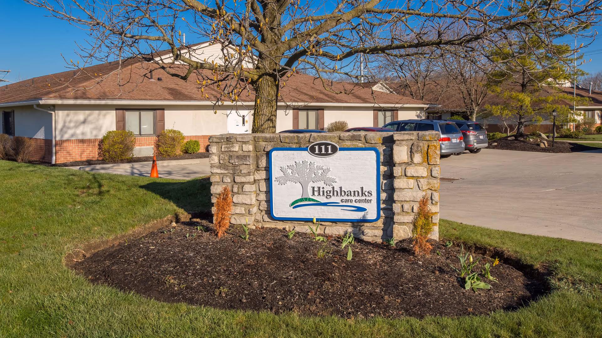Stone sign with the number 111 and the text 'Highbanks care center' in front of a single-story building with a brown roof and beige walls, surrounded by grass, small bushes, and a parking lot with several cars under a clear blue sky.
