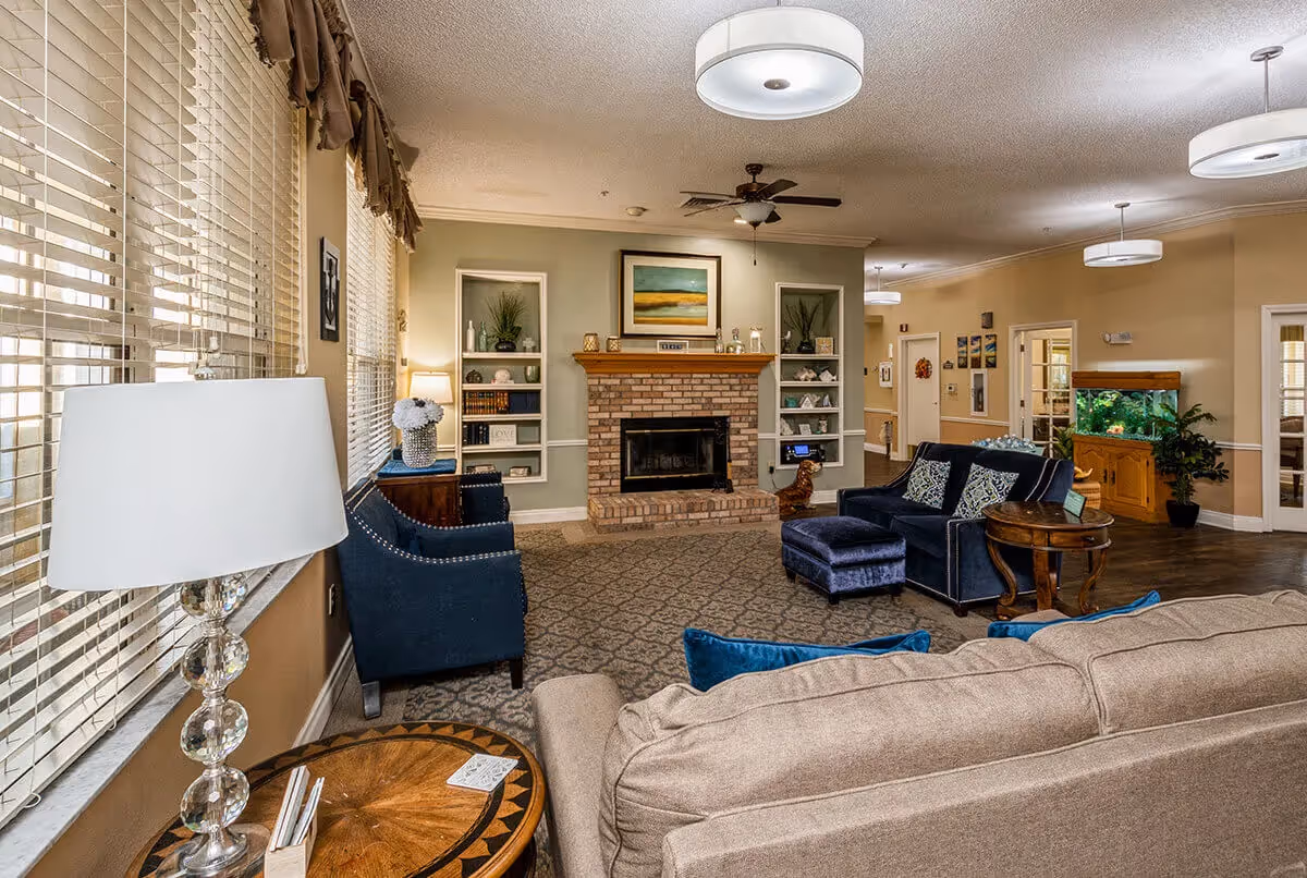 A cozy living room area with a beige sofa, blue armchairs, and a matching ottoman arranged around a brick fireplace. The fireplace is flanked by built-in shelves decorated with books, plants, and ornaments. Large windows with blinds and valances let in natural light. The room features a patterned carpet, ceiling fans, and modern light fixtures. In the background, there is a wooden cabinet with a large fish tank and some wall art.