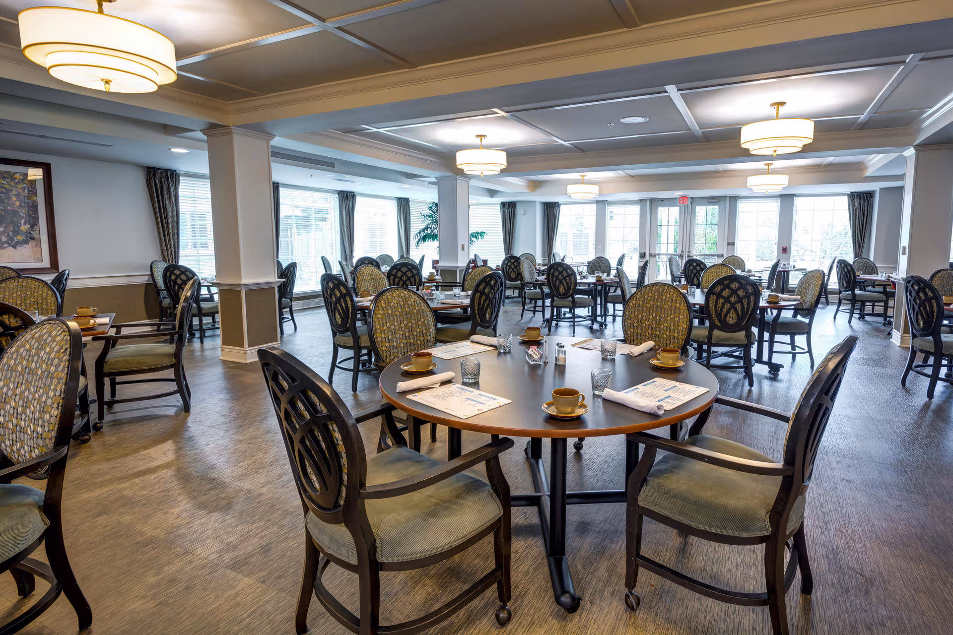 A spacious dining room in a senior living facility with multiple round tables and chairs arranged neatly. Each table is set with cups, glasses, napkins, and menus. The room has large windows with curtains allowing natural light to fill the space, and ceiling lights provide additional illumination. The floor has a wood-like finish, and the overall atmosphere is clean and inviting.