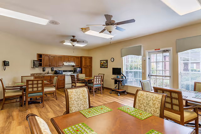 A bright and spacious dining area in a senior living facility with several wooden tables and chairs. The room features a kitchen area with wooden cabinets, a stove, and a microwave. There are ceiling fans and large windows letting in natural light, with a door leading outside.