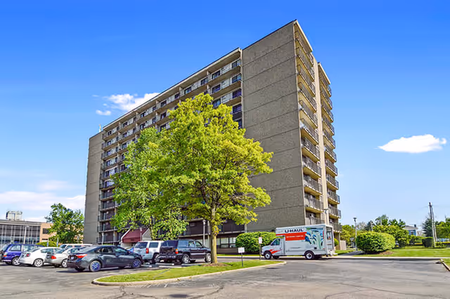 Exterior view of a multi-story senior living facility building with balconies, surrounded by a parking lot with several parked cars and a U-Haul truck, under a clear blue sky.