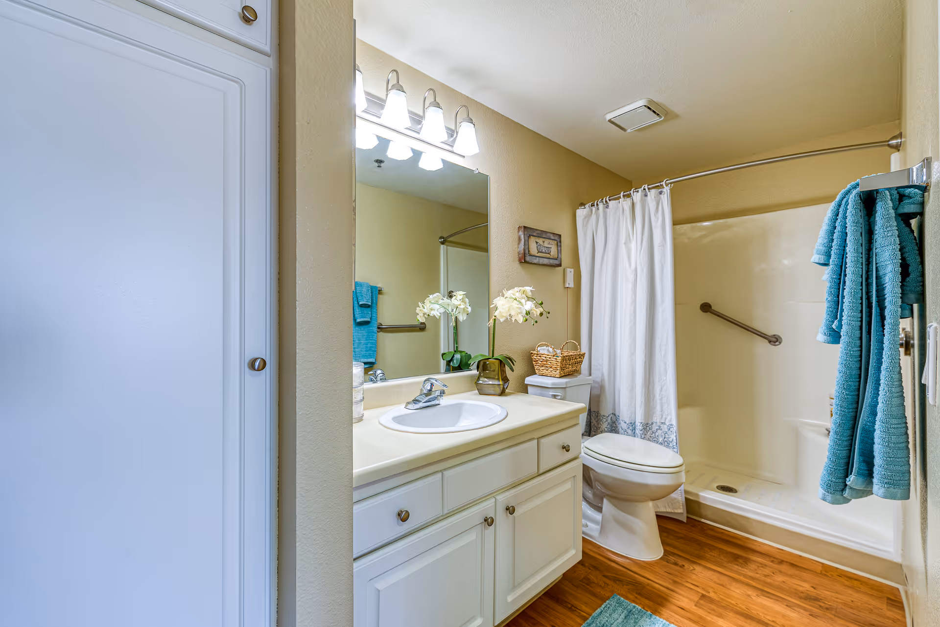 A clean and well-lit bathroom featuring a white vanity with a sink, a large mirror with four light fixtures above it, a toilet with a wicker basket on top, and a shower area with a white curtain and grab bar. Blue towels hang on a rack next to the shower, and the floor has a wood-like finish.